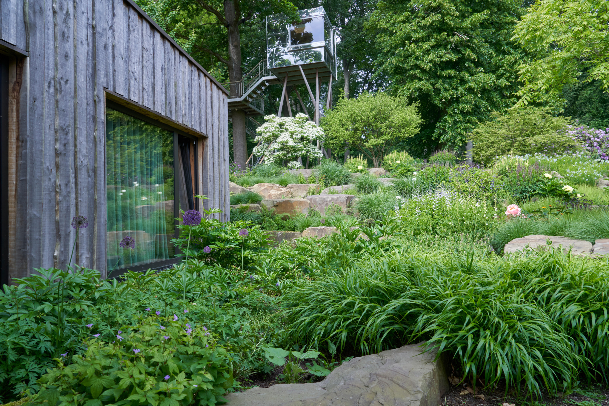 Een tuin op een helling met stenen bij een houten huis, op de achtergrond een uitzichtpunt