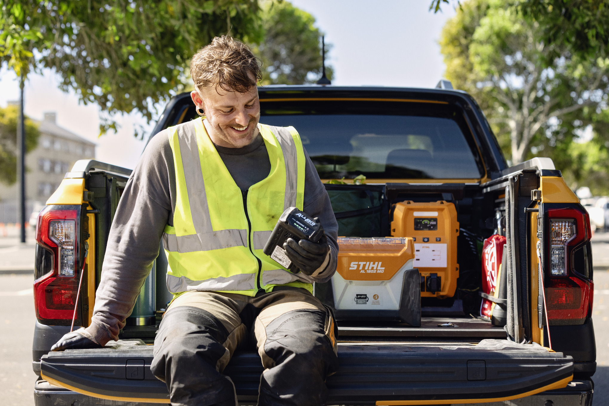 De zitting van de mens op de achterklep van een pick-upvrachtwagen die het ladingsniveau van zijn ALLPRO-batterij controleert. Op de achtergrond staan het snellaadapparaat STIHL AL 1802 MO en de mobiele krachtcentrale PS 3000