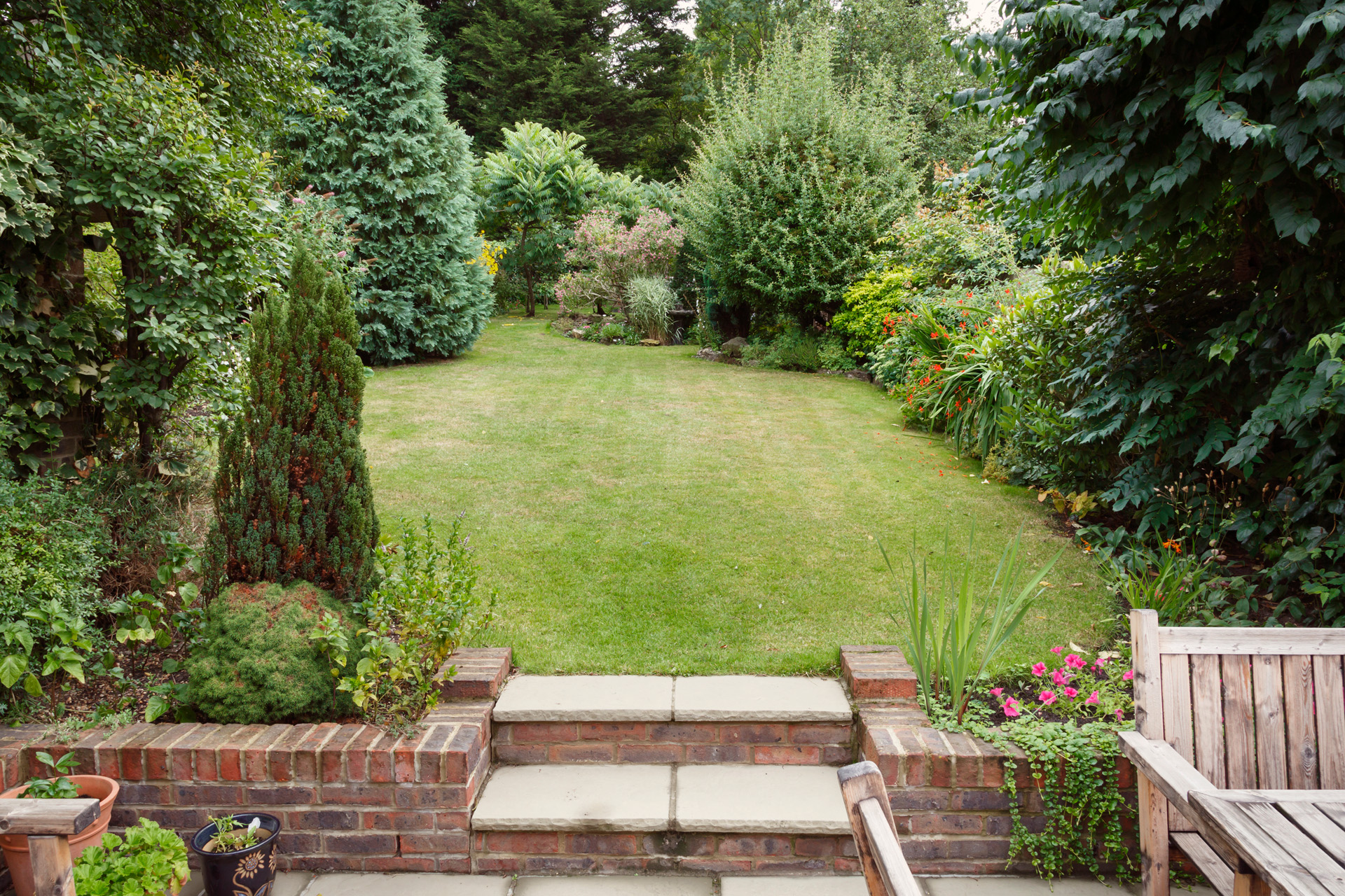 Kleine tuin met terras, tuinmeubelen en plantenbakken op de voorgrond, daarachter grasveld met boompjes en struiken.