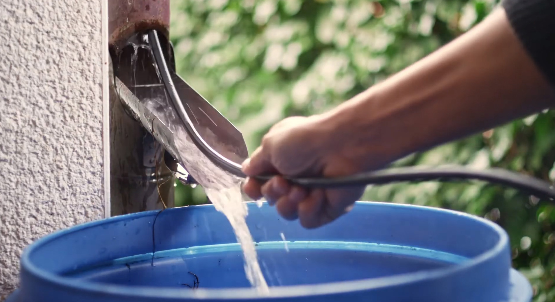 Een flexibele, metalen stang wordt omhoog geduwd in de regenpijp terwijl water in een blauwe regenton stroomt