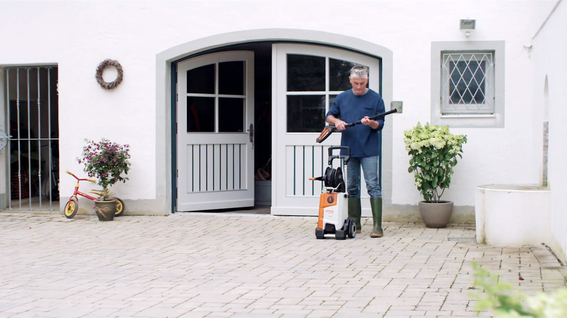 Een man die voor een wit huis staat, bereidt zich voor om de gevel te reinigen met een STIHL hogedrukreiniger