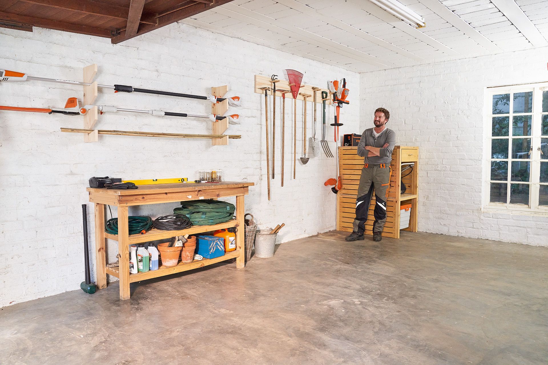 Een man staat met gekruiste armen naast een zelfgemaakte tuingereedschaphouder in een garage 
