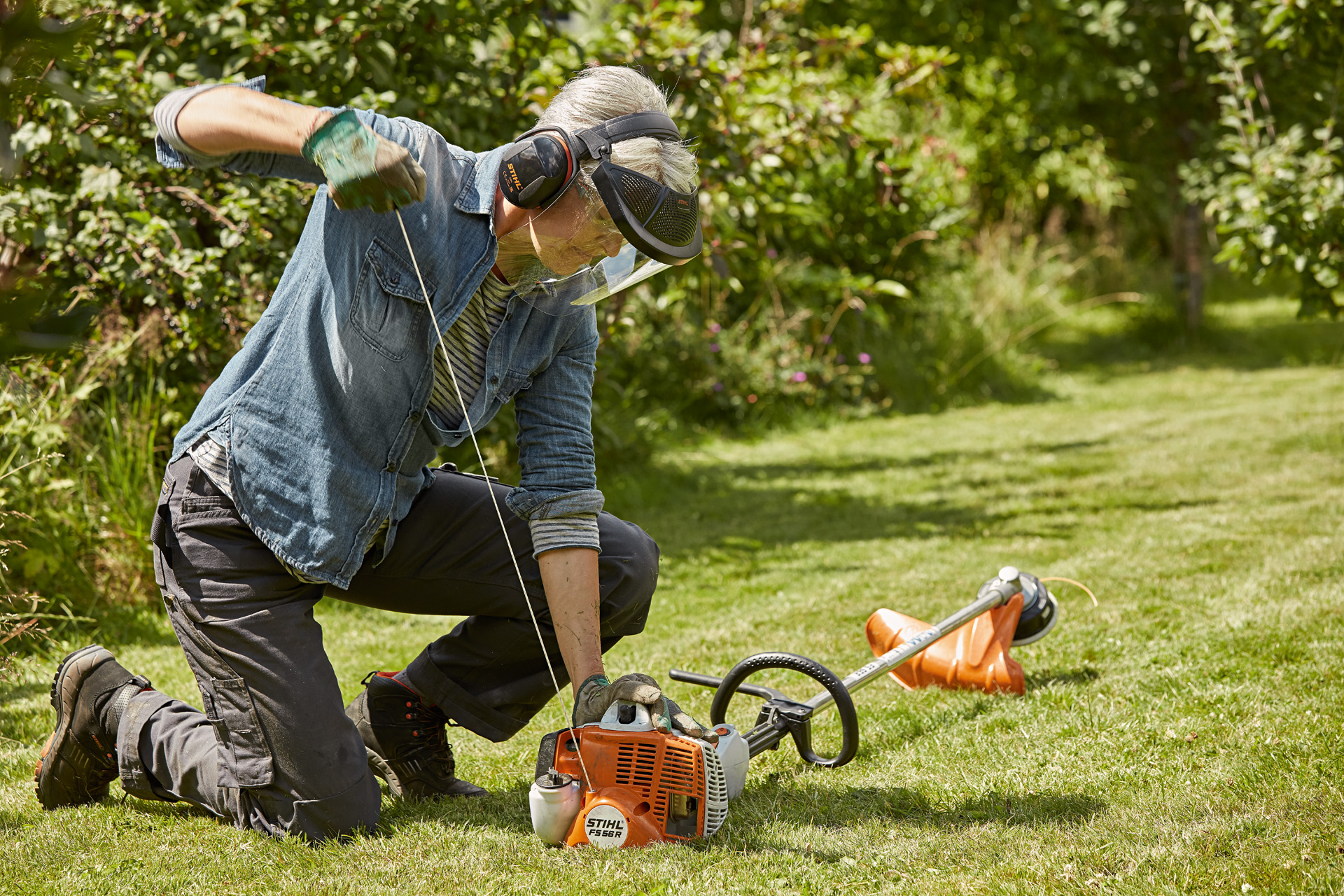 Een vrouw met gehoor- en gelaatsbescherming start een STIHL FS 56 R benzine-kantenmaaier op een begroeid terrein.