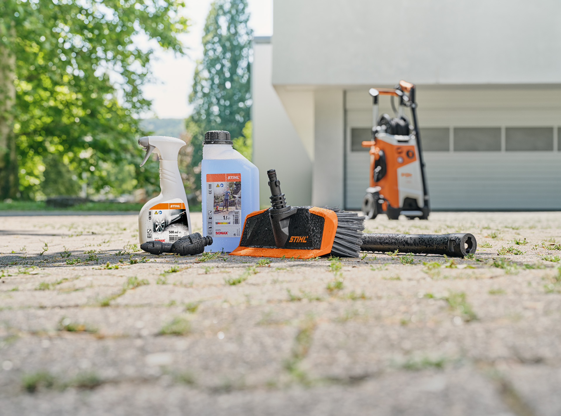  STIHL velgenreiniger, autoshampoo en wasborstel op de grond vóór de garage. Op de achtergrond een STIHL hogedrukreiniger