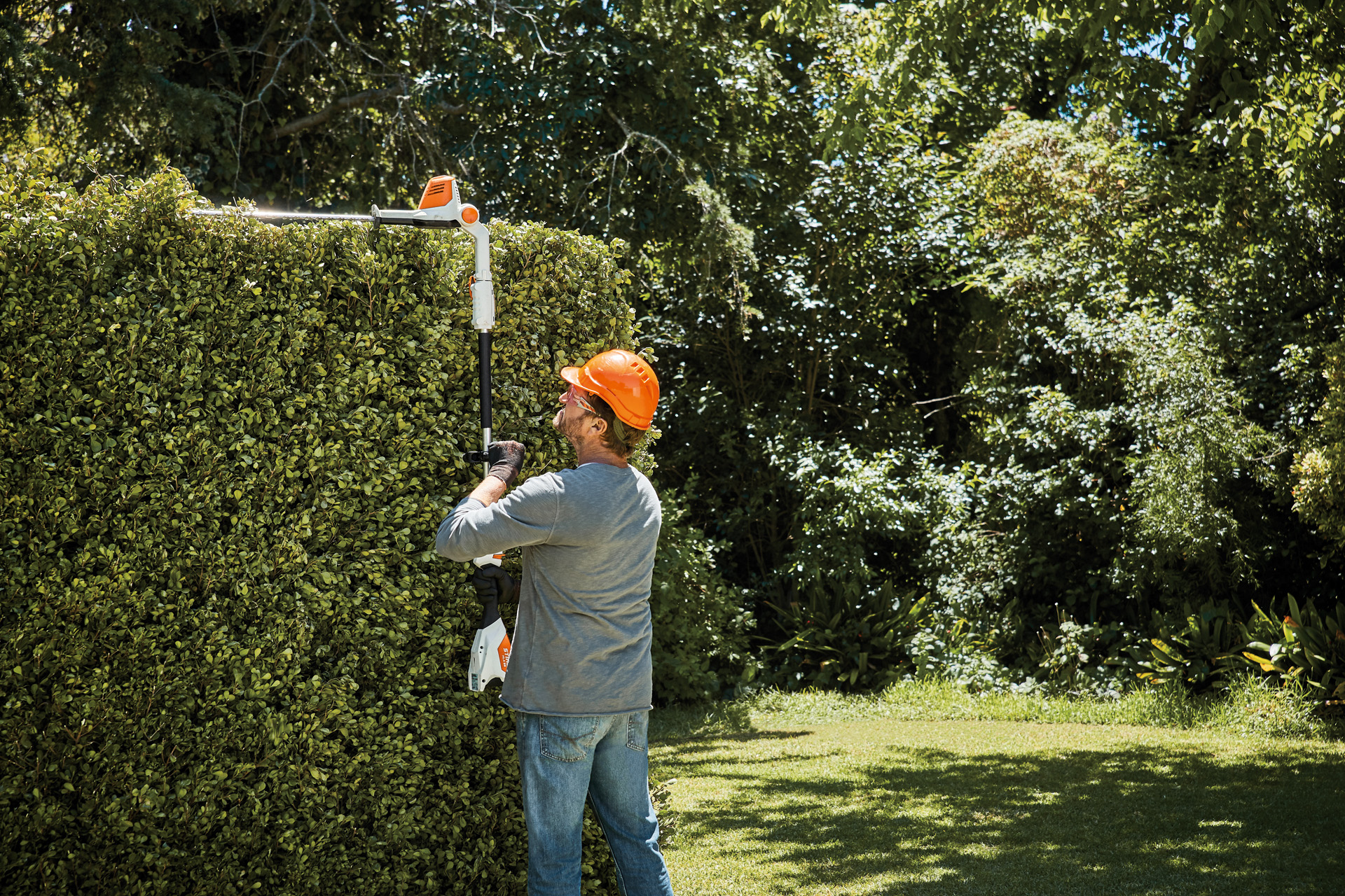 Een man snoeit de bovenkant van een hoge heg met behulp van de STIHL HLA 56 heggenschaar op steel