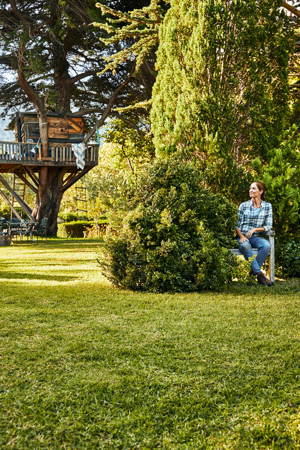  Een vrouw zit op een bankje in een tuin met struiken en gazon, met een boomhut op de achtergrond.