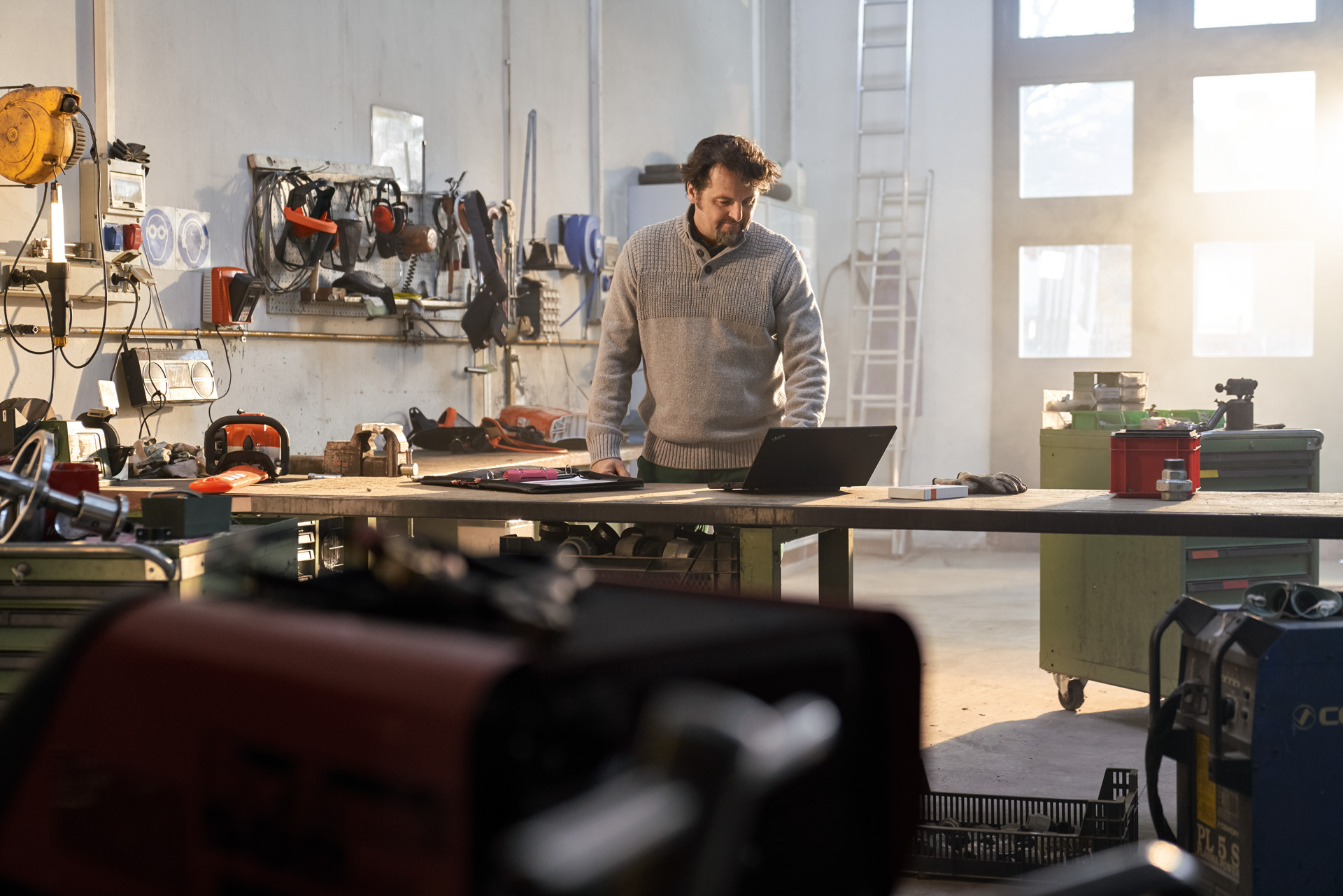 Mann in grauem Pullover steht in einer Werkstatt vor einem Laptop