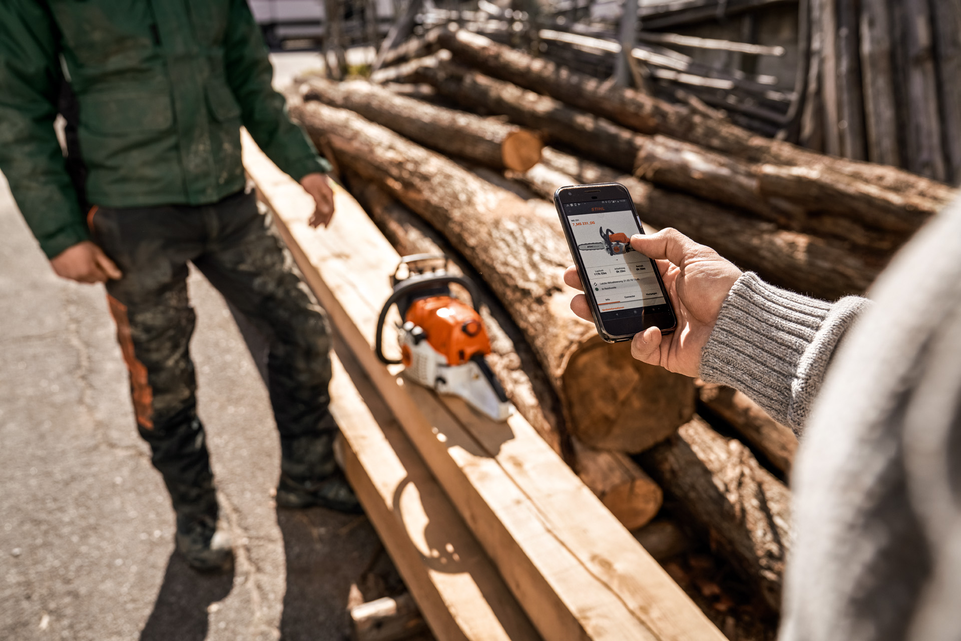 STIHL connected App wordt gebruikt op een smartphone, met een kettingzaag op een stapel hout op de achtergrond
