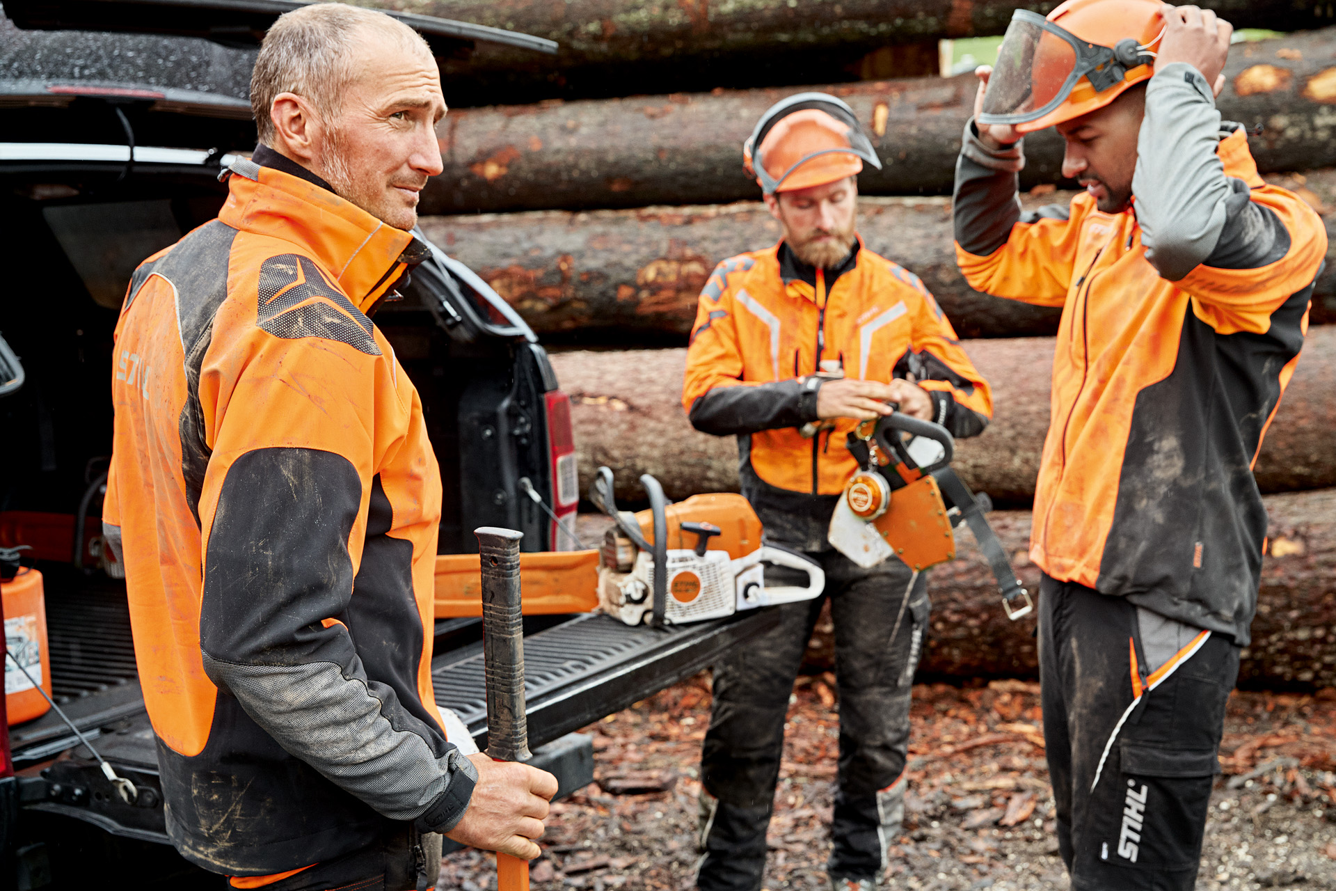 Drie mannen in STIHL veiligheidskleding staan voor een stapel hout en bereiden zich voor om te werken met een STIHL kettingzaag