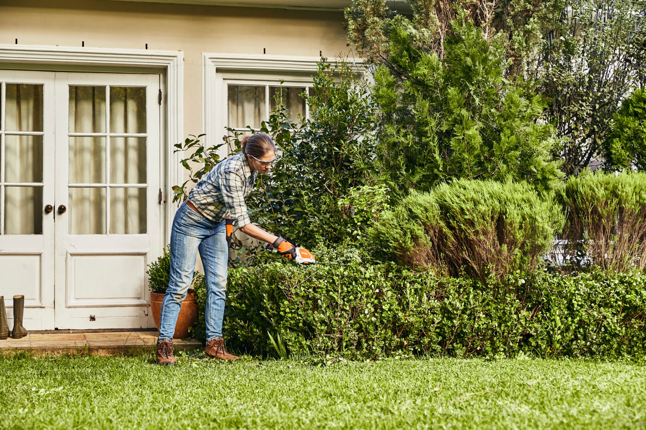  Vrouw snoeit buxusheggen voor een wit huis met een STIHL HSA 26 accugras- en buxusschaar