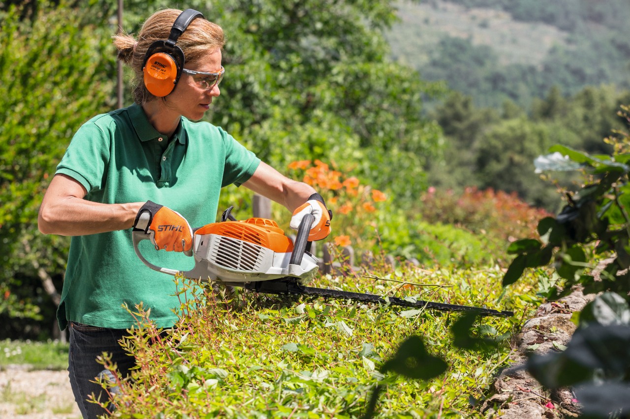 Vrouw met gehoorbescherming en veiligheidsbril snoeit met een STIHL HS 46 benzineheggenschaar een heg